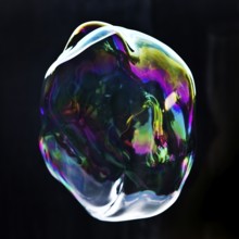 A large iridescent and floating soap bubble in the colours of the rainbow against a black