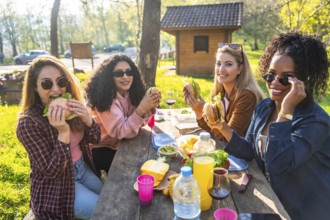 Four young women enjoying a picnic lunch outdoors, eating hamburgers and sharing good times