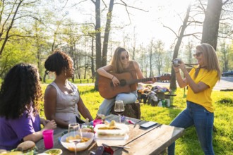Four female friends enjoying a picnic in a park, with one playing guitar, another taking photos,