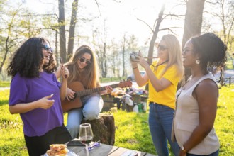 Multi ethnic women having fun at picnic, playing guitar, taking pictures and singing together