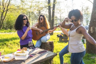 Diverse women enjoying a lively picnic in a park, singing joyfully, playing guitar, and dancing