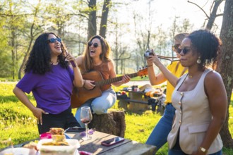 Four female friends enjoying a picnic in a park, singing, playing guitar, taking photos, and having