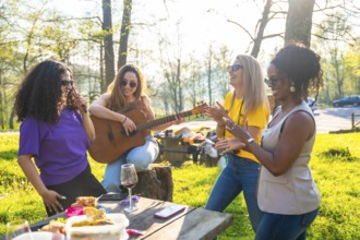 Four diverse female friends are enjoying a picnic in a park, singing, playing guitar, and clapping