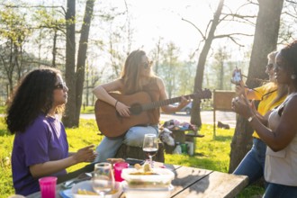 Group of friends enjoys a picnic in a park, with one playing guitar while another takes photos
