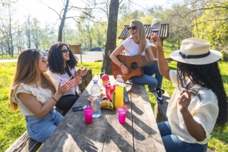 Four diverse women are having a picnic in a park, playing guitar, clapping, drinking wine, and