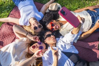 Four cheerful female friends are lying on a blanket in a park, taking a selfie with a smartphone