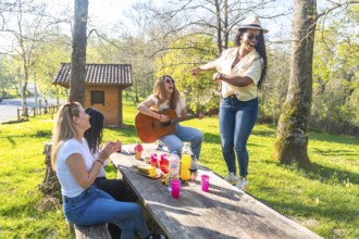 Four friends enjoying a picnic in a park, with one playing guitar, another dancing, and the others