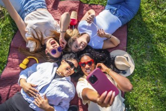 Four female friends are relaxing on a blanket in a park, taking a selfie with a smartphone on a