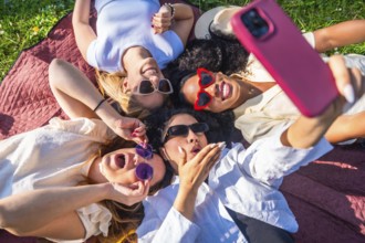 Four cheerful young women wearing sunglasses are lying on a blanket in a park, taking a selfie with