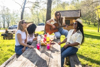 Four female friends are sitting at a wooden table, playing guitar and enjoying a picnic outdoors,