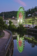 A large Ferris wheel glows in green and red and is reflected in a river at dusk, 950 years of Calw,