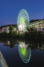 A red illuminated Ferris wheel with reflection in the river against a city backdrop in the evening,