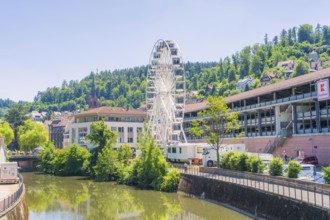 A white Ferris wheel against an urban backdrop and a green, wooded hill in bright sunlight, 950