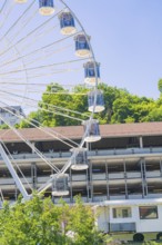 A large Ferris wheel towers over a building, surrounded by green trees under a clear blue sky, 950