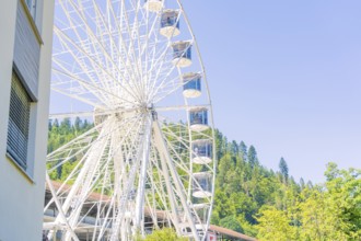 A large Ferris wheel stands next to a building with a wooded hill and clear sky in the background,