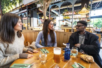 Three friends enjoying drinks and engaging in lively conversation at a stylish wooden table in a