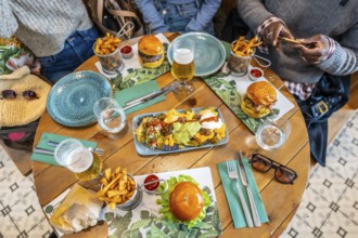 Overhead view of friends sharing a meal of burgers, nachos, french fries, and drinks at a wooden