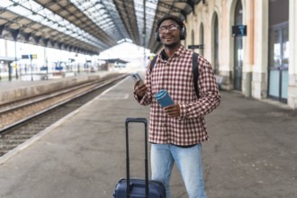 Young black male tourist waiting for his train while listening to music and holding a reusable