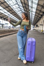Tourist taking selfie with smartphone while waiting for train arrival at station platform, holding