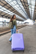 Tourist wearing headphones walking in train station pulling a purple suitcase and carrying a