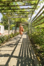 Woman in a pavilion, covered by leaves, with long shadows on the ground, surrounded by summer