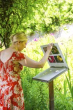 A woman looks at an information board in the middle of a green and flowery landscape, Botanical