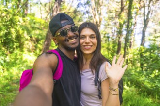 Interracial couple smiling and waving while taking a selfie during a vibrant hike in a lush, green