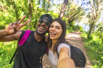 Interracial couple wearing backpacks taking a selfie while hiking in a lush forest, enjoying their