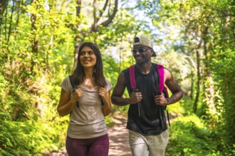 Interracial couple wearing backpacks walking in a forest trail, enjoying the beautiful nature