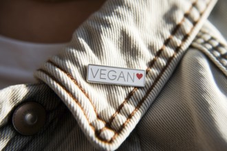 Close-up of silver pin with VEGAN text and red heart on worn cotton jacket. Symbol of ethical