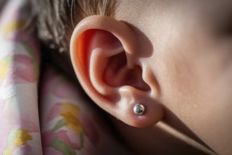 Delicate silver earring stud in baby's ear bathed in warm golden light. Tender moment with subtle