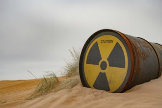 Rusty radiation barrel half-buried in sand under overcast sky. Symbol of environmental danger.