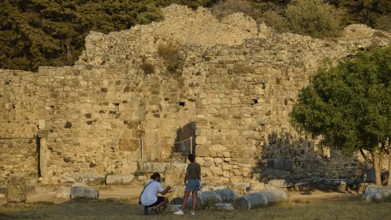 Two people in front of an old stone wall in a setting surrounded by trees, Roman bathhouse,