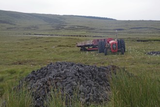Old red tractor standing in boggy fields against a hilly backdrop with grey sky, peat cutting, Isle