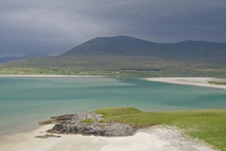 Wide sandy beach meets turquoise waters, surrounded by green hills and mountains under a gloomy
