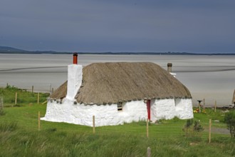 White thatched house on a green meadow against a sandy coastal background under a blue sky,
