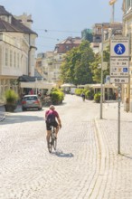 A cyclist rides along a sunny city street surrounded by historic buildings and trees, Merano, Italy