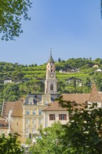 City skyline with a striking church tower surrounded by green hills against a clear sky, Merano,