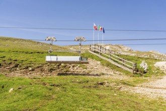 Chairlift in front of mountain landscape with flags and green slope under clear blue sky, Alpe di