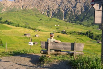 A person sits on a bench and looks out over an expansive mountain landscape with rolling hills,