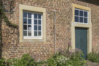 A rustic brick wall with two windows, wooden door and flowering plants, Haus Welbergen, Ochtrup,