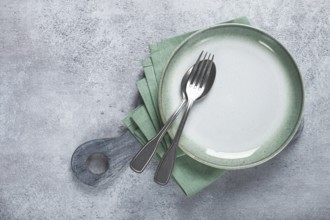 An empty plate and cutlery laid out, on a cutting board, with a green napkin, a gray table, top