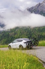 White car with bicycles on road, surrounded by misty mountain landscape and green forest, electric