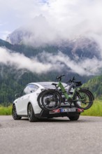 A car with bicycles drives along a road against a dramatic mountain backdrop and clouds, electric