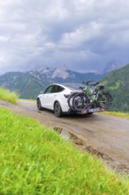 White car with bicycles on mountainous road, surrounded by green landscape and cloudy sky, electric