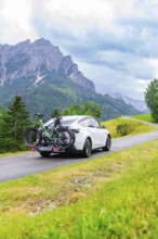 White car with bicycles on a road in a mountainous, green landscape with cloudy sky, electric car