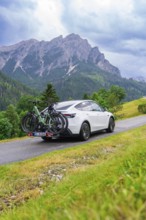 White car with bicycles on road in green, mountainous surroundings under cloudy sky, electric car