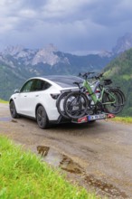 White car with bicycles on wet road, surrounded by mountains and rain clouds, electric car on