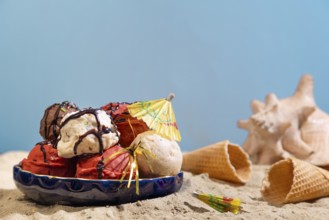 Various flavours of ice cream with chocolate sauce and parasol on sand with shells