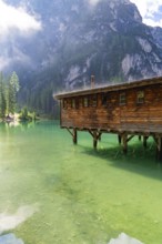 A wooden house stands on stilts above a clear, green mountain lake, surrounded by tall trees and
